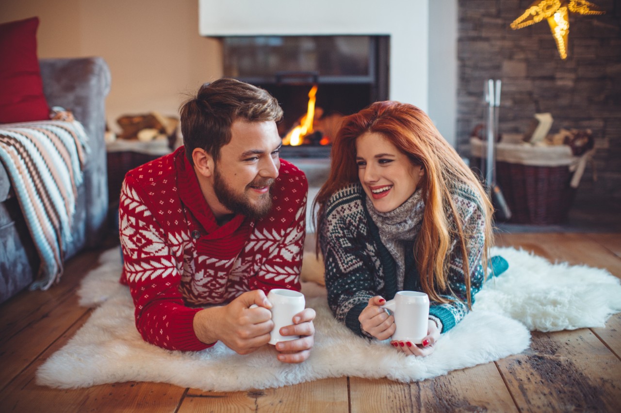 Couple on vacation at mountain cabin. Lying on the floor on a blanket and drinking hot drink by a fireplace in a cozy  living room on Christmas. Wearing festive knitted sweaters. Austrian Alps.